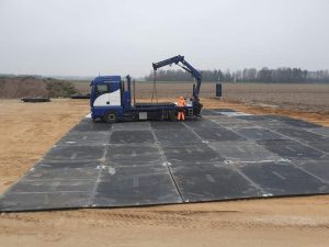 Ciężarówka z podnośnikiem na placu budowy, otoczona ziemią i pustym polem, widoczni pracownicy.