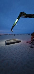 Dźwig na plaży w nocy, oświetlający placuszki drewna na piasku, w tle widoczne łodzie i miasto.