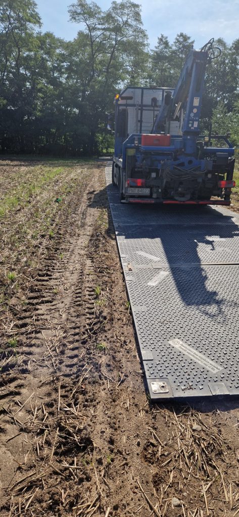 Maszyna rolnicza na polu z widocznym pasem z płyt metalowych i śladami gąsienic na ziemi. W tle drzewa.