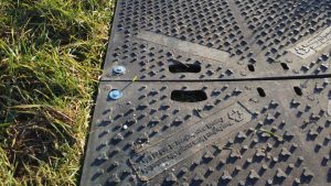 Fragment powierzchni z gumowej maty, widoczne śruby, otwory, elementy oznaczeń oraz trawa w tle.