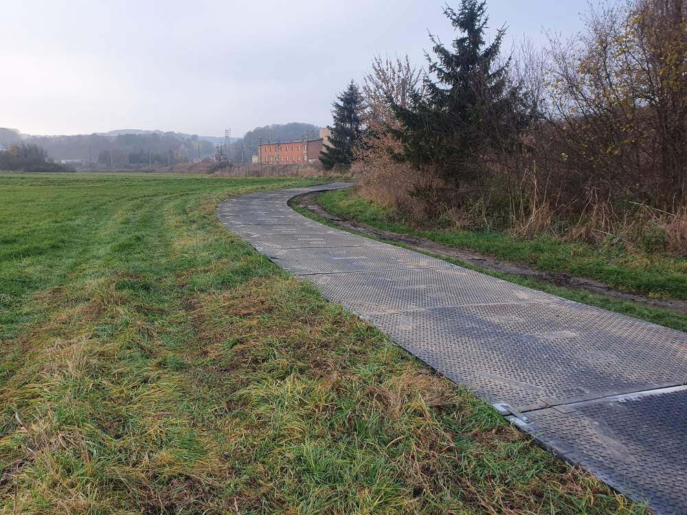 Wyboista ścieżka prowadzi przez zieleń, z drzewami po bokach i budynkiem w tle.