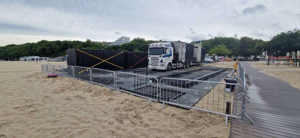Na plaży stoi ciężarówka obok kontenera, otoczona barierkami. W tle drzewa i zabudowania.
