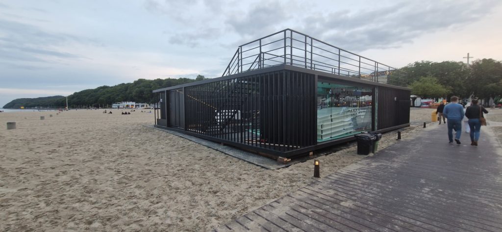 Nowoczesny budynek na plaży, oszklony, z tarasem i ogrodzeniem. W tle piaskowa plaża i zieleń.