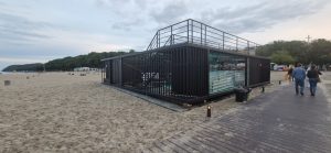 Nowoczesny budynek na plaży, oszklony, z tarasem i ogrodzeniem. W tle piaskowa plaża i zieleń.