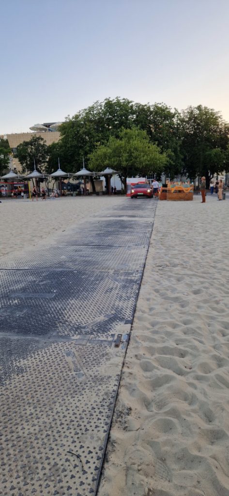 Ścieżka na plaży prowadząca do strefy z zielenią i budkami, w tle widać piasek i ludzi.