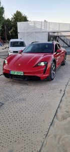 Czerwony samochód Porsche Taycan zaparkowany na placu, obok stoi biały pojazd. W tle widoczne elementy infrastruktury.