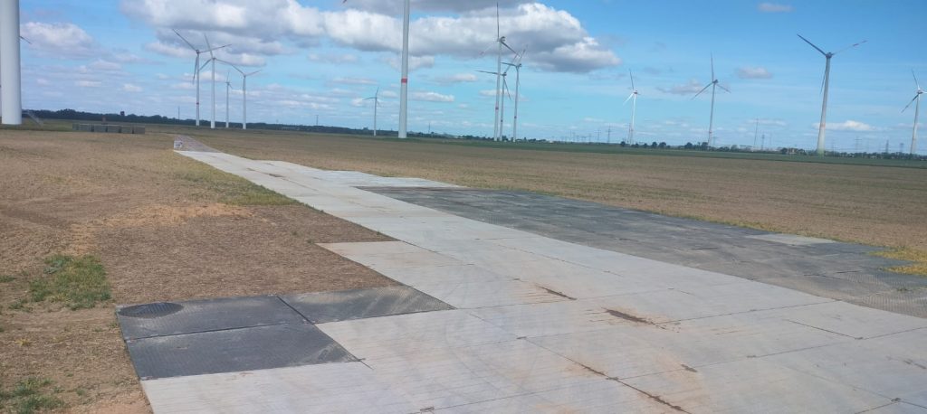 Widok na szeroką, asfaltową drogę w polu, z turbinami wiatrowymi w tle pod jasnym niebem.