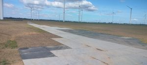 Widok na szeroką, asfaltową drogę w polu, z turbinami wiatrowymi w tle pod jasnym niebem.