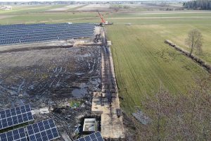 Widok z góry na pole słoneczne z panelami, obok teren budowy i zielone pola. W tle widoczne krajobrazy wiejskie.