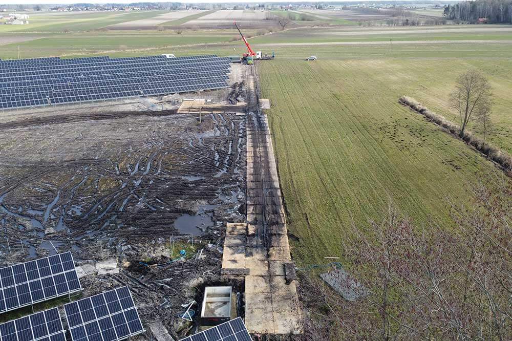 Widok z góry na pole słoneczne z panelami, obok teren budowy i zielone pola. W tle widoczne krajobrazy wiejskie.