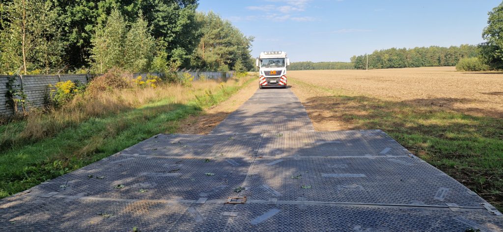 Droga prowadząca przez pole, z metalową platformą i pojazdem służbowym na końcu. W tle widok na zadrzewiony teren.