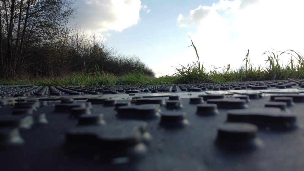 Widok na drogę z bliska, z wyraźnym wzorem powierzchni, otoczoną trawą i drzewami w tle. Niebieskie niebo z chmurami.