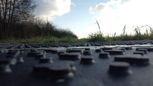 Widok na drogę z bliska, z wyraźnym wzorem powierzchni, otoczoną trawą i drzewami w tle. Niebieskie niebo z chmurami.