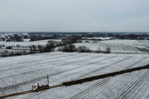 Widok na zimowy krajobraz z pola pokrytego śniegiem oraz ciężarówką na drodze. W tle zarys wsi.