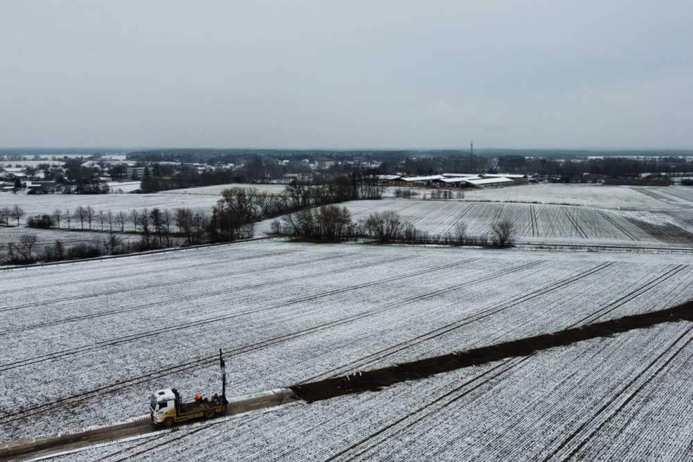 Widok na zimowy krajobraz z pola pokrytego śniegiem oraz ciężarówką na drodze. W tle zarys wsi.