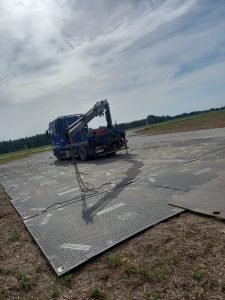 Tir z podnośnikiem na metalowej nawierzchni w wiejskim terenie pod jasnym niebem.