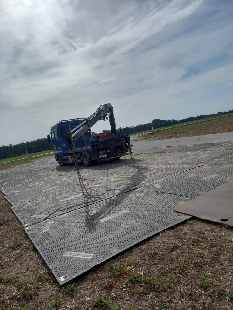 Tir z podnośnikiem na metalowej nawierzchni w wiejskim terenie pod jasnym niebem.