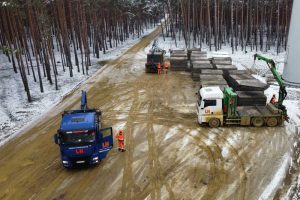 Trucks i pracownicy w odblaskowych kamizelkach na błotnistej drodze w lesie, obok składowane materiały.