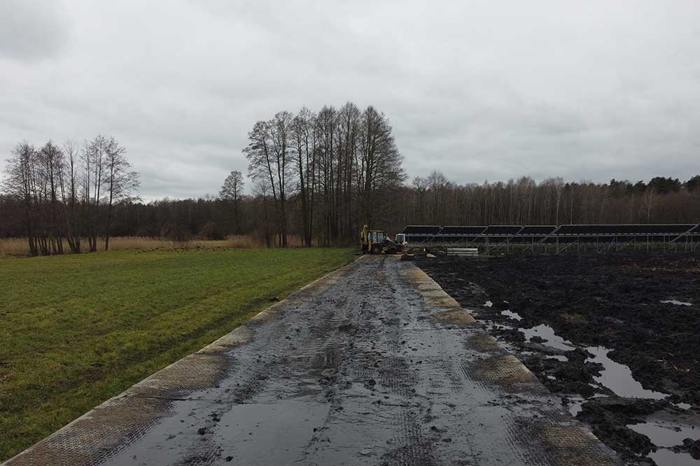 Droga prowadząca przez teren z płytami, z widokiem na solary i otaczające drzewa. Ziemia mokra i błotnista.