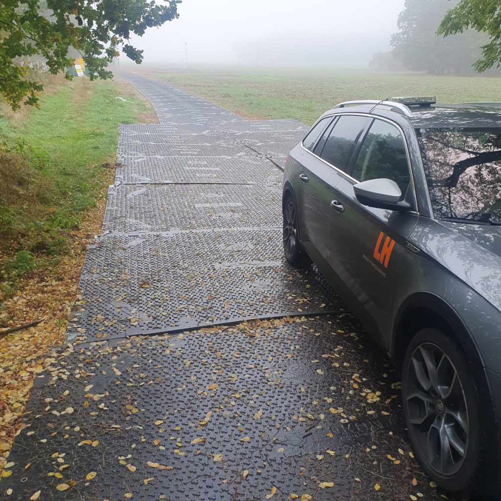 Na zdjęciu widać szary samochód zaparkowany na pokrytej liśćmi drodze w mglistym otoczeniu.