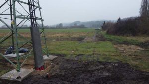 Obiekt radiowy na polu, z widoczną betonową podstawą i wytyczonym terenem, w tle zalesione wzgórza.