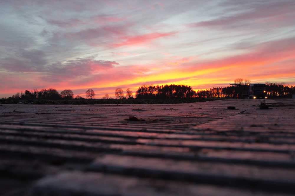 Zachód słońca nad polami, z intensywnymi odcieniami różu i pomarańczu oraz zarysami drzew w tle.