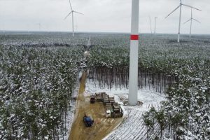 Plac budowy w lesie, z wiatrakami w tle, pokryty śniegiem, na drodze widoczne materiały budowlane.