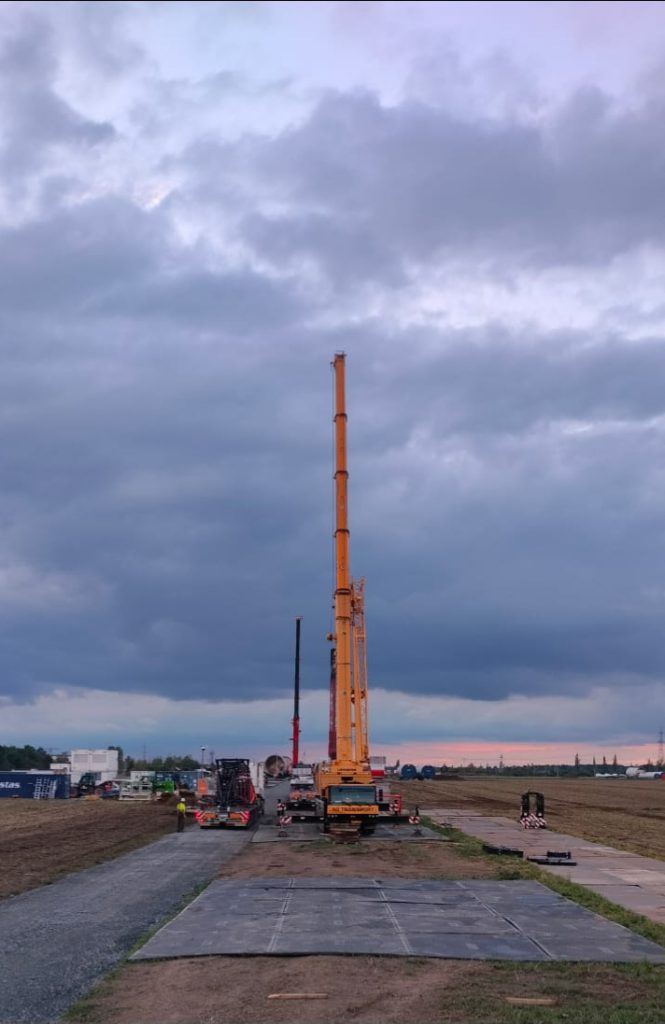 Wysoka żuraw budowlany ustawiony na polu, z chmurami w tle. Widać także sprzęt budowlany i pracowników.