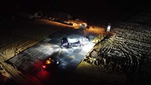 Prace budowlane w nocy na placu budowy, oświetlona maszyna i pojazdy na działce z ziemią.