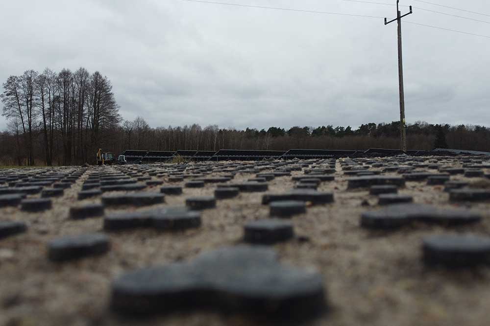 Widok na teren z panelami słonecznymi, w tle drzewa i słup energetyczny, pochmurna pogoda.