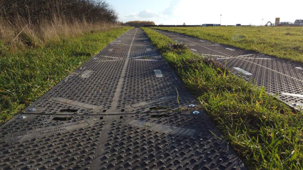 Ścieżka z metalowych płyt na tle zielonej łąki, z rosnącą trawą po obu stronach. W tle widoczne niebo.