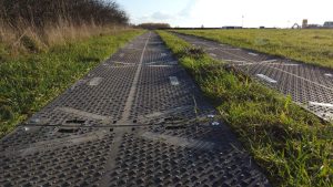 Ścieżka z metalowych płyt na tle zielonej łąki, z rosnącą trawą po obu stronach. W tle widoczne niebo.