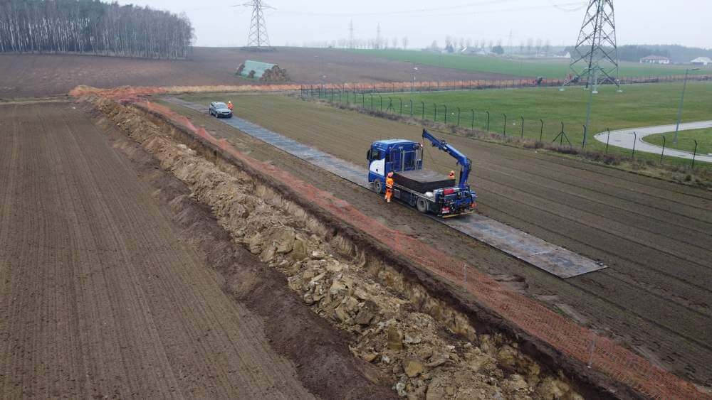 Na zdjęciu jest ciężarówka budowlana i droga na polu. W tle widać tereny rolnicze i wysokie słupy energetyczne.