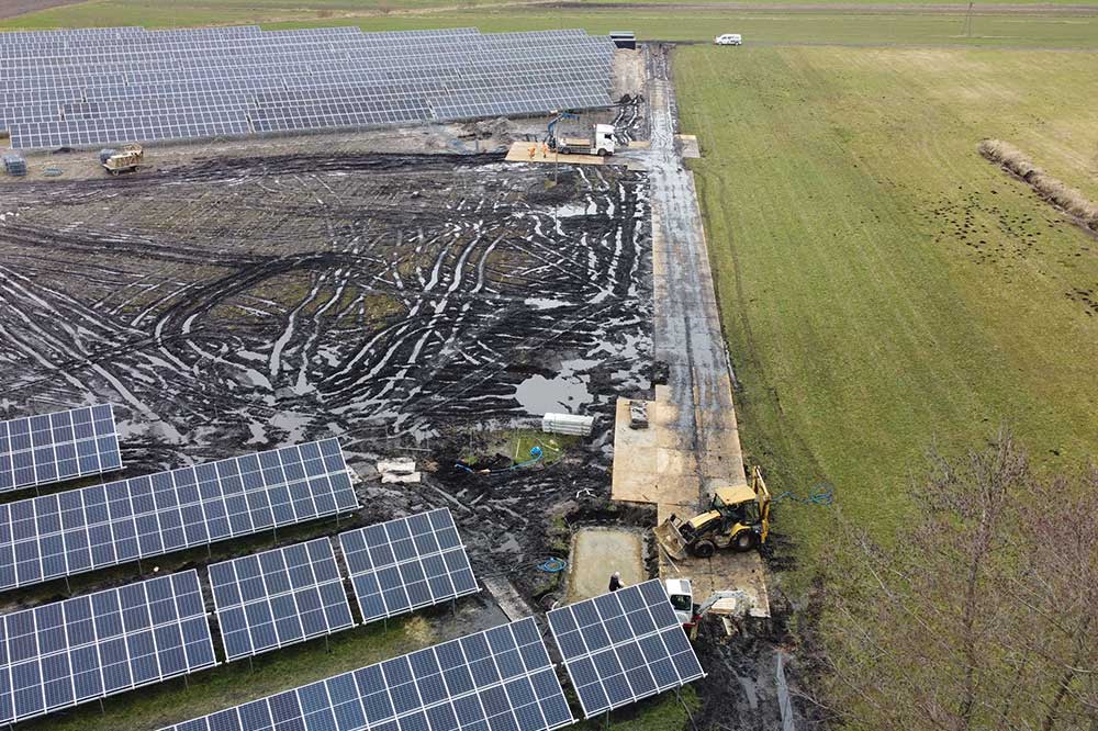 Widok z góry na pole paneli słonecznych, z widocznymi śladami na ziemi i pojazdami budowlanymi w tle.