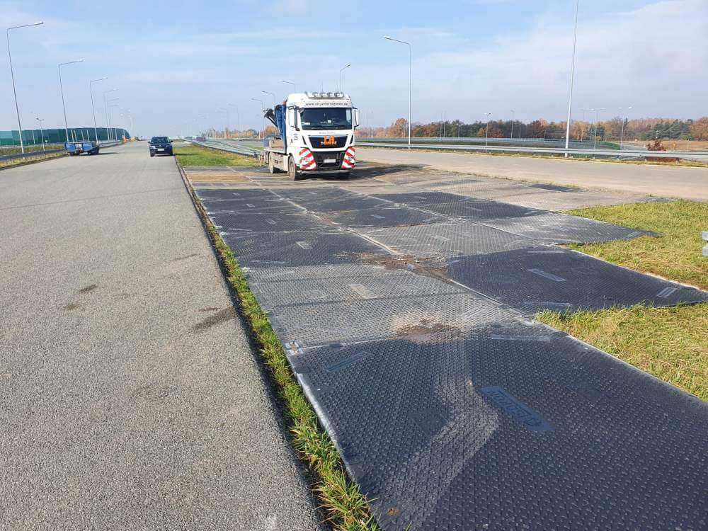 Ciężarówka na drodze obok ułożonych płyt, w tle widoczna jezdnia i elementy krajobrazu.