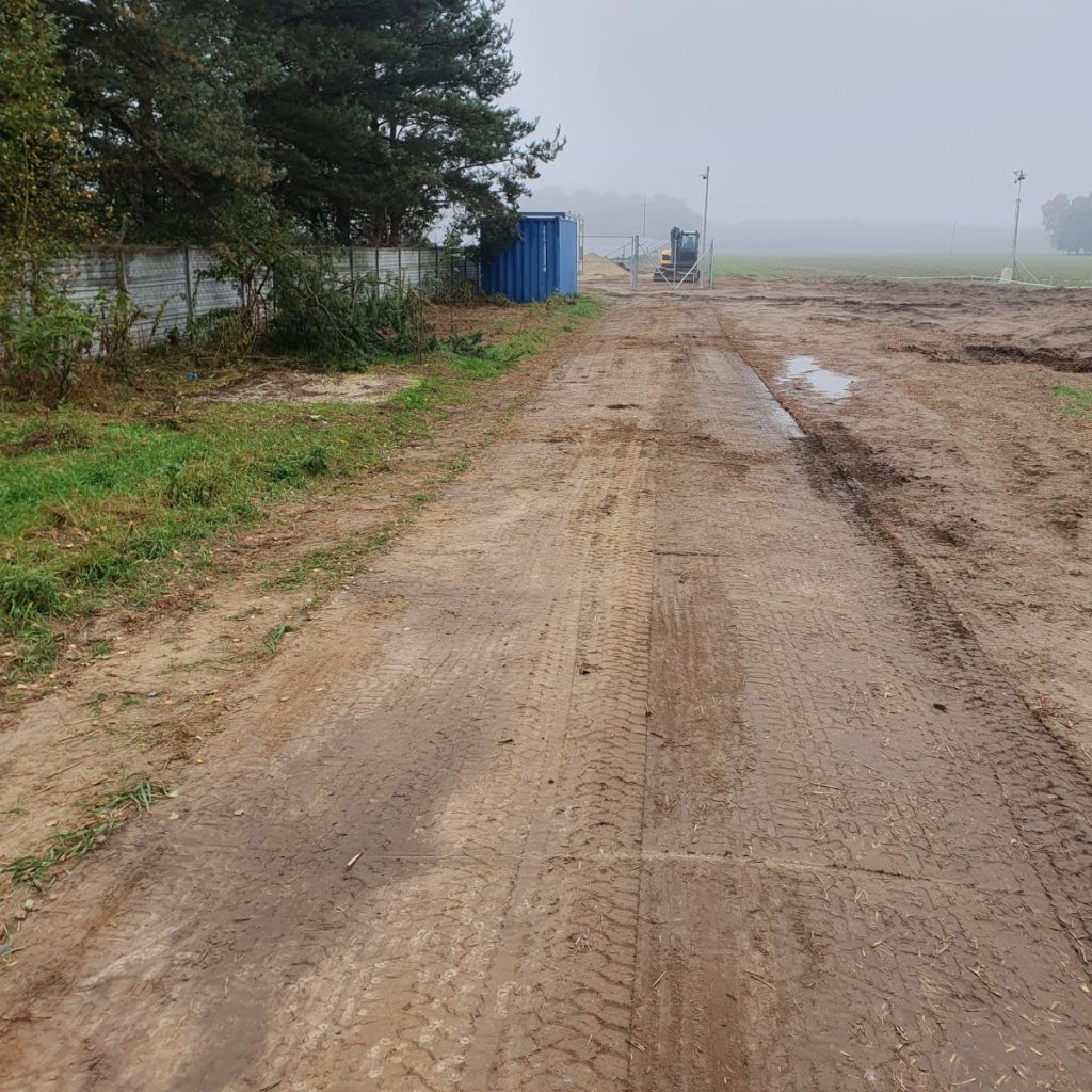 Błotnista droga na terenie zarośniętym drzewami, w tle kontener i maszyny budowlane, mglista pogoda.