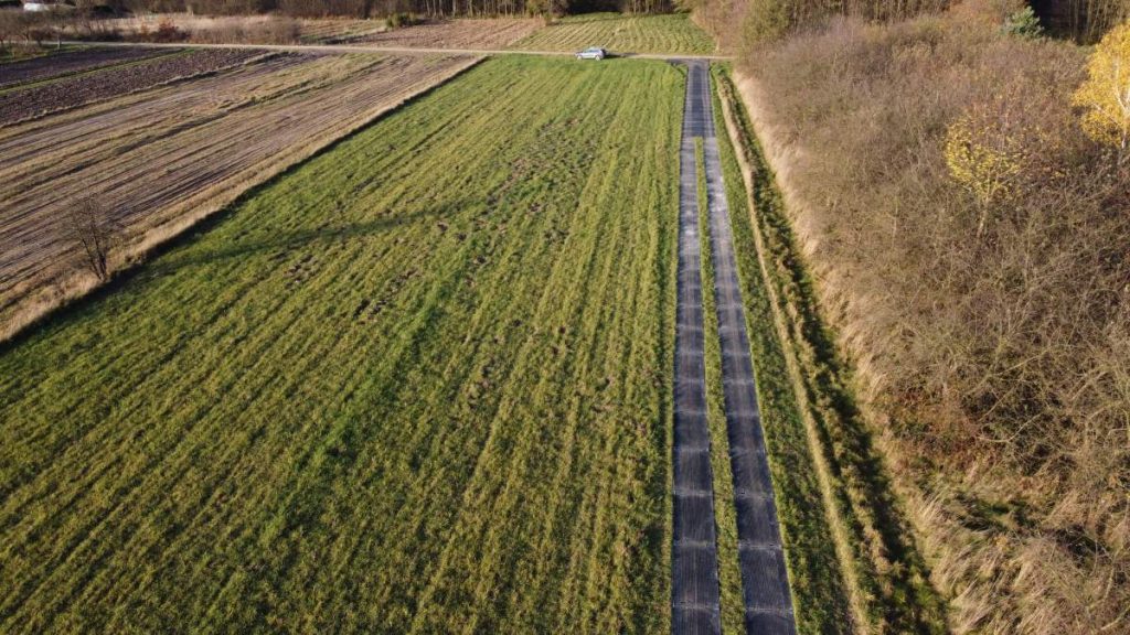 Droga prowadząca przez pole, po obu stronach otoczone zaroślami, z widokiem na las w tle. W oddali widać samochód.