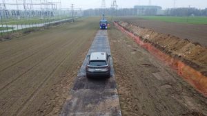 Na zdjęciu widać drogę poprowadzoną przez pole, z samochodem na środku i pojazdem służbowym w tle.