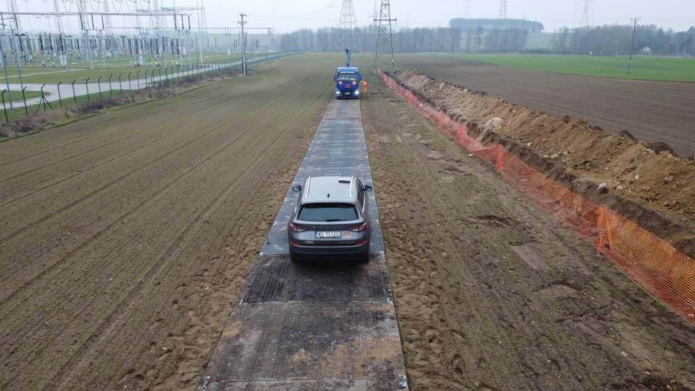 Na zdjęciu widać drogę poprowadzoną przez pole, z samochodem na środku i pojazdem służbowym w tle.