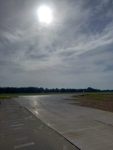 Widok na pusto zamkniętą drogę na lotnisku, otoczoną zielenią, z szarym niebem i slyszącym słońcem.