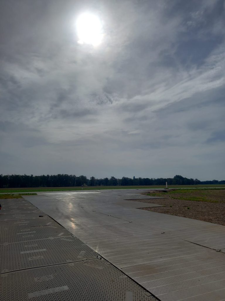 Widok na pusto zamkniętą drogę na lotnisku, otoczoną zielenią, z szarym niebem i slyszącym słońcem.