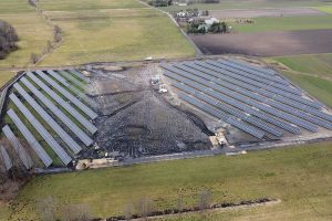 Pole słoneczne z panelami fotowoltaicznymi na dużej powierzchni, otoczone zielonymi terenami i drogą.