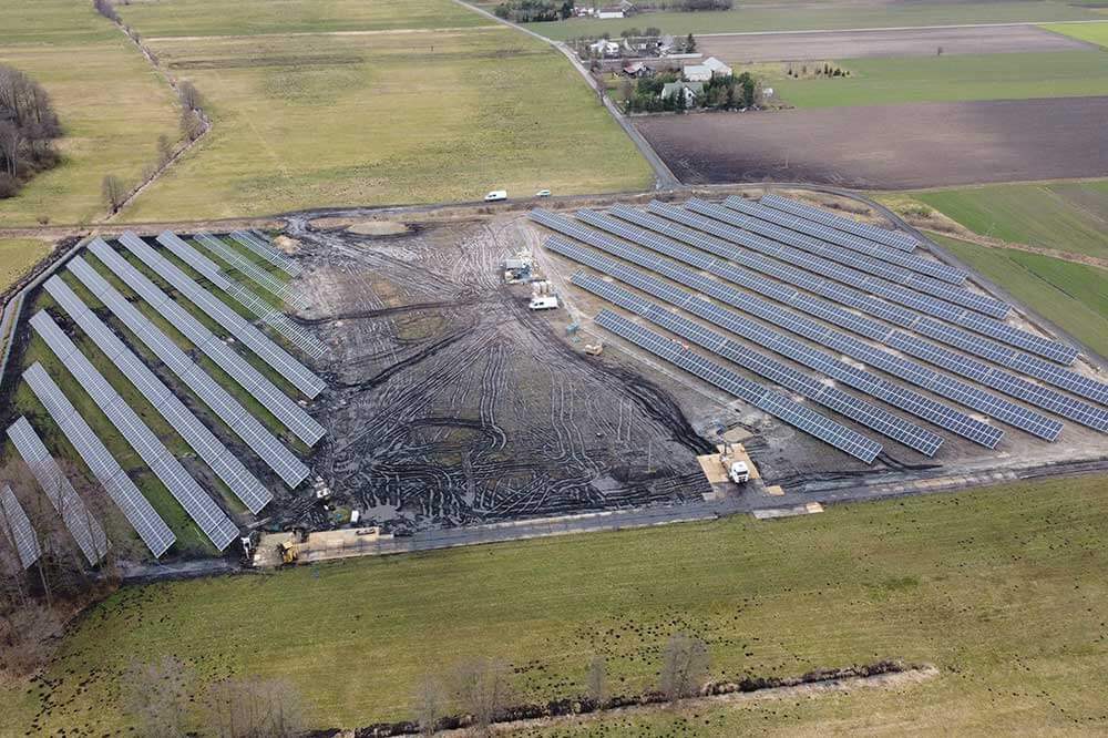 Pole słoneczne z panelami fotowoltaicznymi na dużej powierzchni, otoczone zielonymi terenami i drogą.