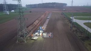 Roboty drogowe na polu, widoczne maszyny, pracownicy w pomarańczowych ubraniach, linie energetyczne w tle.