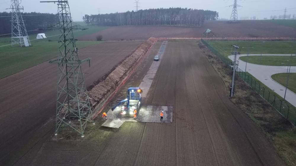 Roboty drogowe na polu, widoczne maszyny, pracownicy w pomarańczowych ubraniach, linie energetyczne w tle.