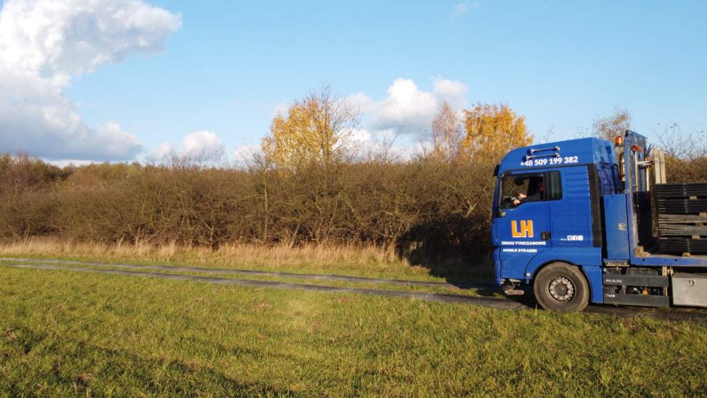 Ciężarówka stoi na drodze przy lesie, w tle widoczne drzewa i błękitne niebo. Złociste liście na drzewach.