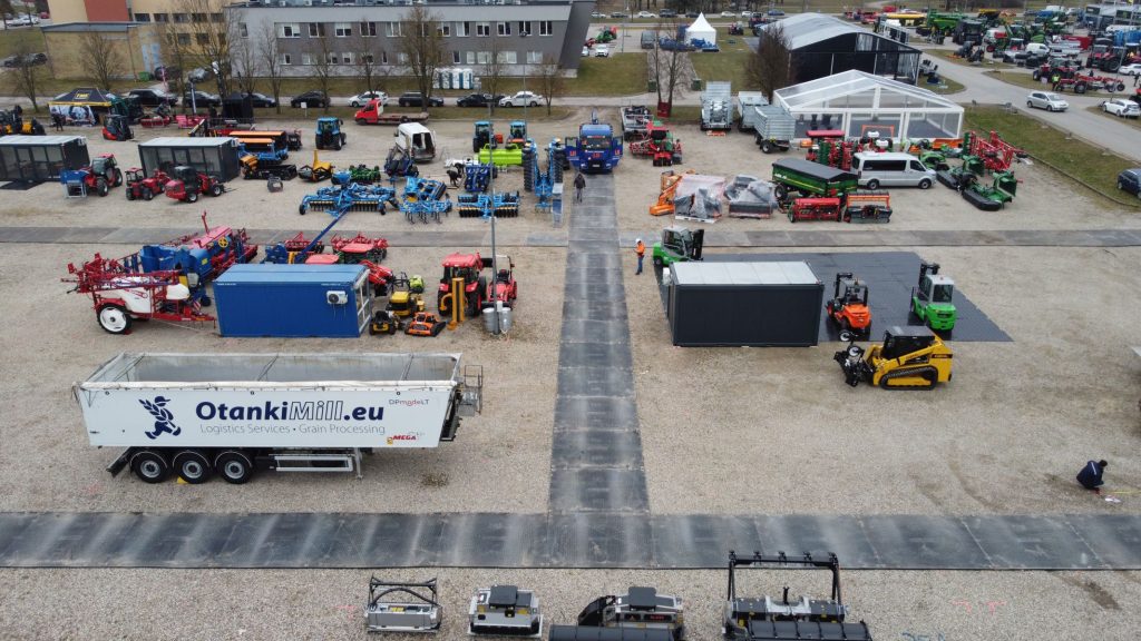 Widok z lotu ptaka na plac z maszynami rolniczymi, przyczepami i kontenerami, otoczony drogami z kostki brukowej.