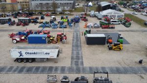 Widok z lotu ptaka na plac z maszynami rolniczymi, przyczepami i kontenerami, otoczony drogami z kostki brukowej.