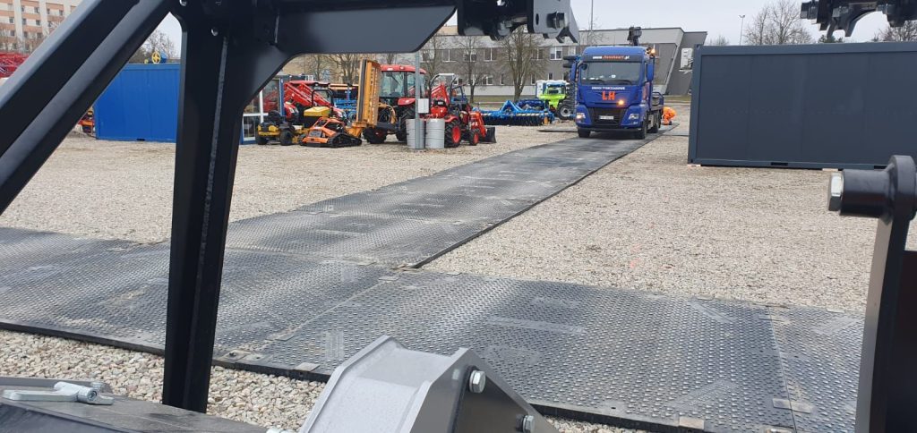 Na obrazie widać plac z maszynami budowlanymi, w tym ciągnikami i ciężarówką na drodze z metalowych płyt.