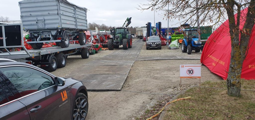 Na zdjęciu widać teren targowy z ciągnikami, maszynami rolniczymi i wozem na naczepie. Obok stoi czerwona namiot.